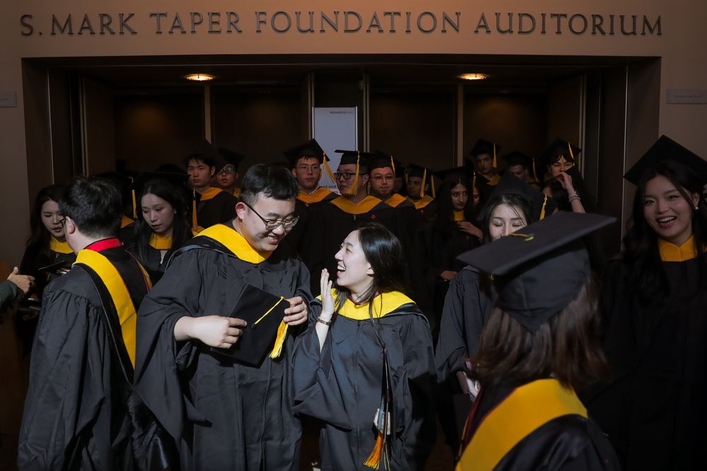 Students dressed in regalia stand in a crowd laughing together. 