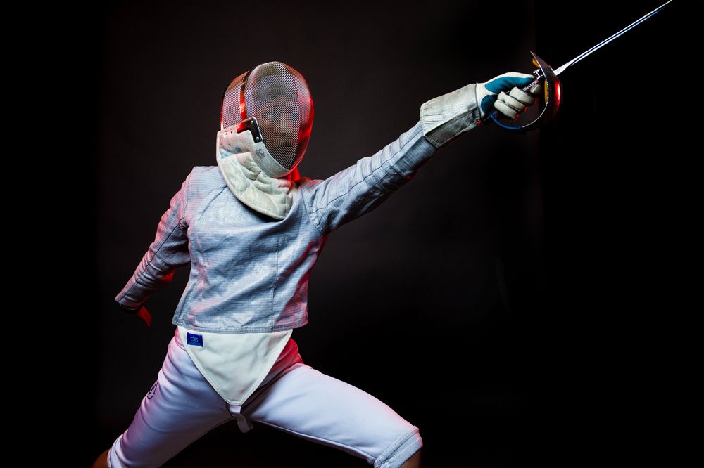A student on the fencing team, dressed in fencing gear, lunges to the side while pointing their sword toward the upper right corner. 