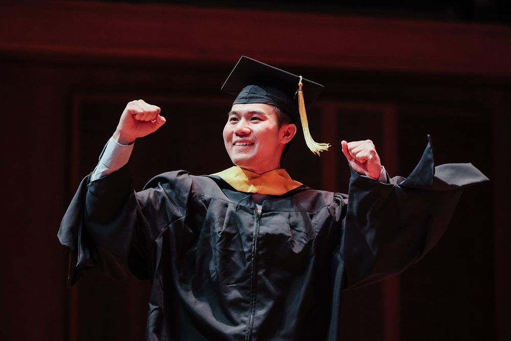 A student wearing regalia cheers with their hands raised in the air. 