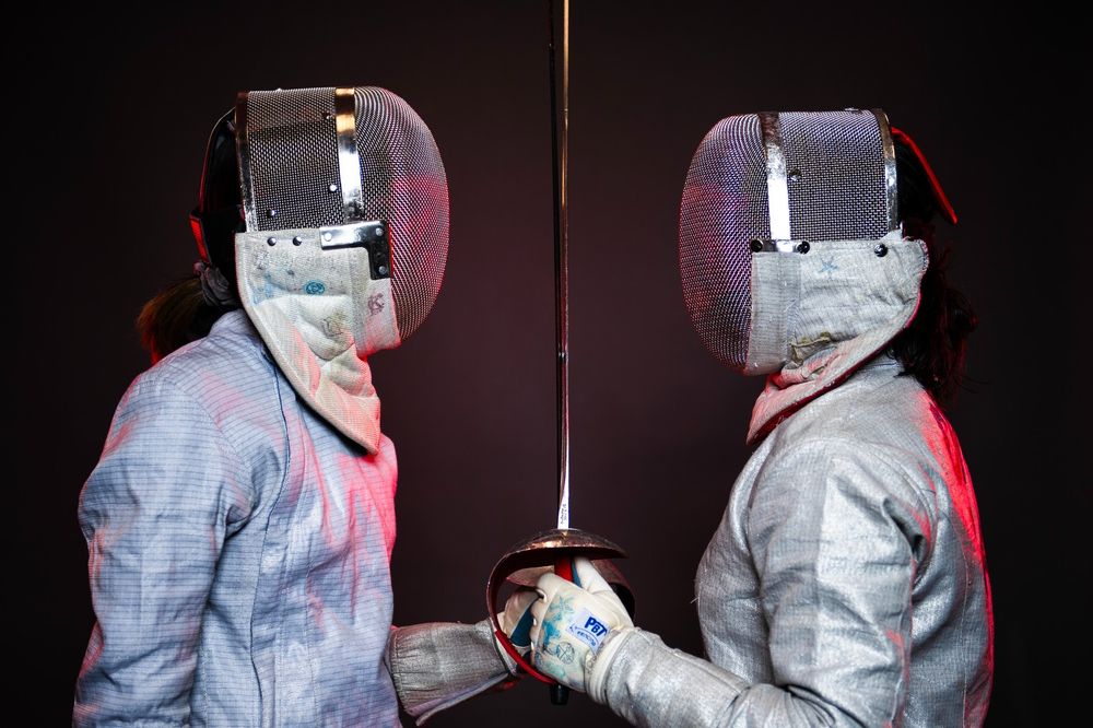 Two students dressed in fencing gear stand facing each other and holding a sword between them. 