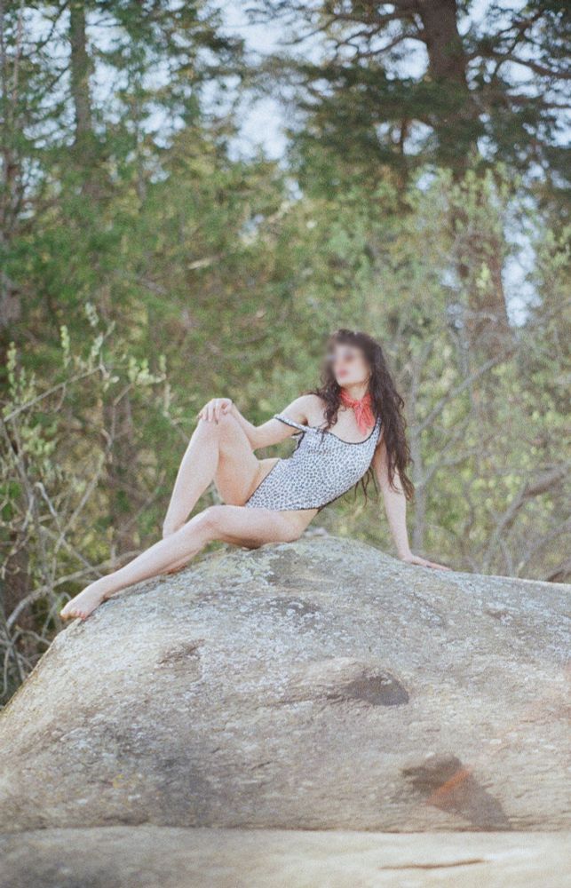 a film photo of sam perched onto of a rock on a beach in the bright sunshine. There is a green forest in her background. She wears a leopard black and white one piece swimsuit and a red scarf around her neck. her long curly black hair is down. She wears sunglasses. She is leaning on the side of her body with her legs outstretched to the side and her gaze looking in the distance