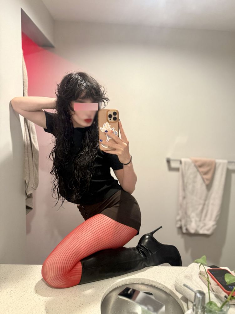a selfie of Sam in the bathroom mirror. Her knee is perched on the counter. The wear bright red tights, knee high black leather stilettos, short brown shorts and a black turtle neck t-shirt. Her dark hair is wet and curly and her lips are red.