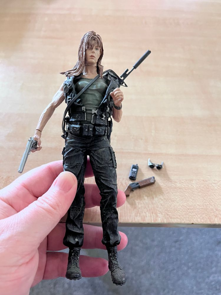 Action figure of Sarah Connor from Terminator 2. She’s in black pants, army green shirt, black tactical vest. She has a handgun in her right hand and a rifle strapped to her back. Accessories displayed on a table are her phone, a sawed off shot gun, and aviator sunglasses. 