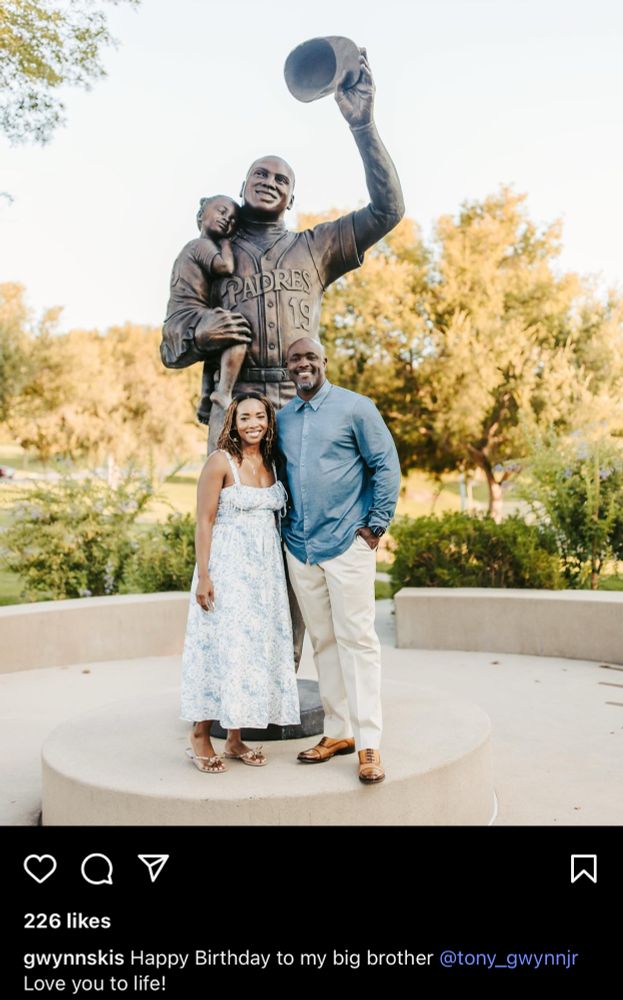 Photo of Tony Gwynn Jr. and Anisha Gwynn Jones standing in front of a statue of their father, Tony Gwynn. Gwynn’s statue depicts him holding Anisha in his right arm as he tips his Padres cap with his left hand.