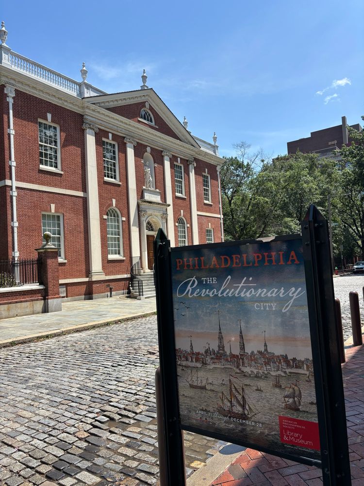 Sign saying “Philadelphia: the Revolutionary City”. Statute of Ben Franklin in the background 