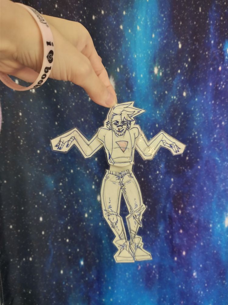 Hand holding up a laminated paper doll of BurakkuHardt's OC Izzy, with moveable ligaments and a messy sketch style. The background is a space-print curtain