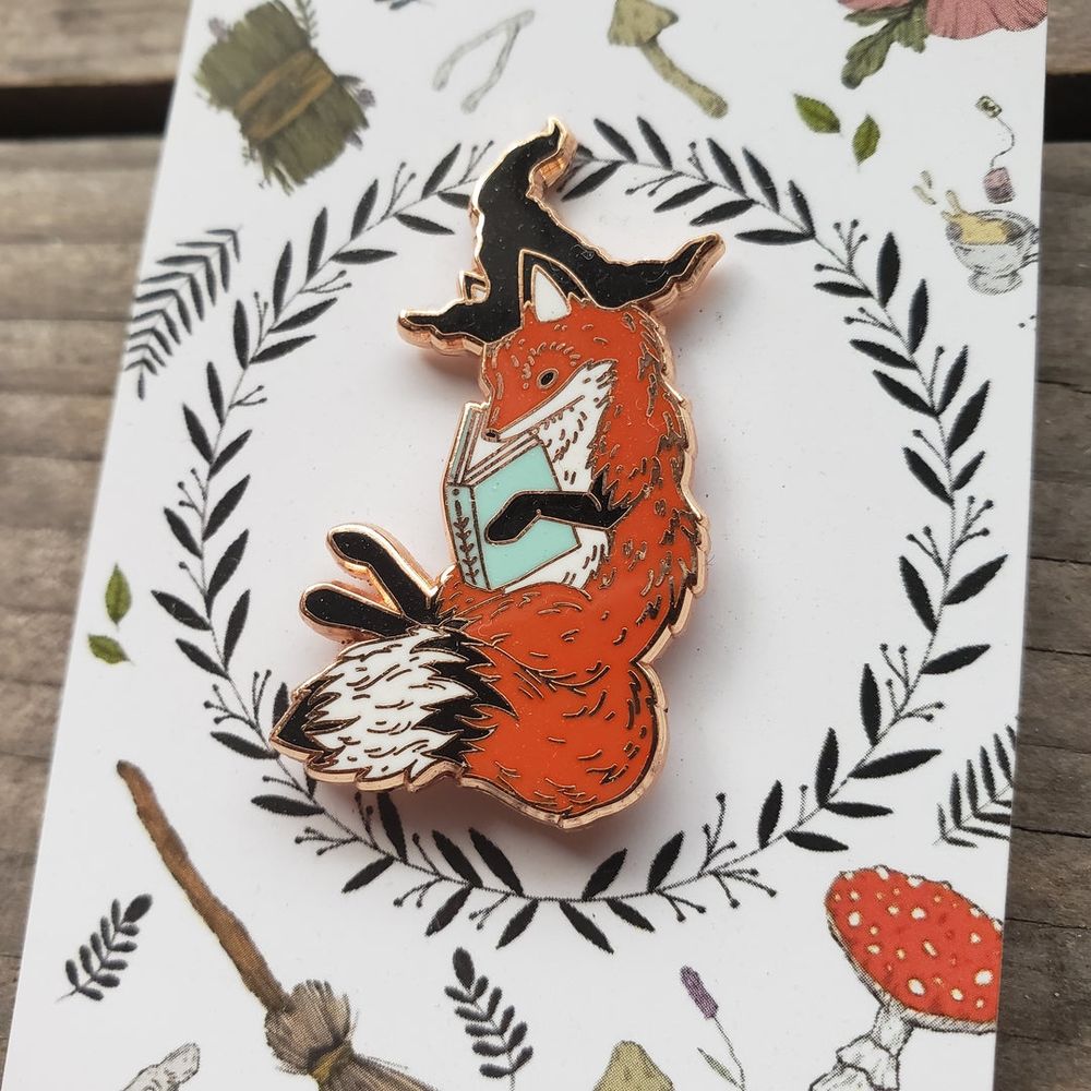 An enamel pin of a red fox, holding a book, and wearing a black witch hat.