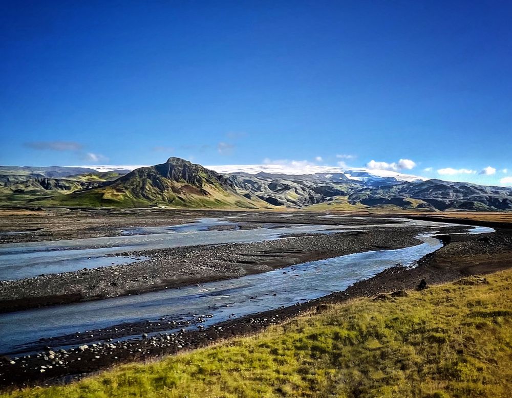 This photo captures a wide and open Icelandic landscape under a bright blue sky. In the foreground, the braided channels of the Hafursá River wind gently through a stony riverbed, reflecting light in shades of silver and pale blue. The riverbanks are a mix of dark volcanic soil and grassy patches in soft green tones.

Beyond the river lies a striking, rugged mountain with steep sides and a greenish-brown hue, set against a backdrop of rolling hills and ridges in varying shades of grey and green. In the distance, the white ice cap of the Mýrdalsjökull glacier stretches across the horizon, blanketing the higher mountains with snow. Hidden beneath that glacier is the powerful and unpredictable volcano Katla. 