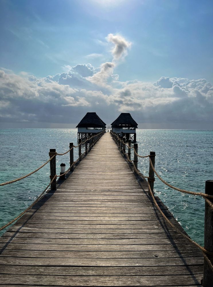 The photo shows a long wooden pier stretching out straight into the turquoise sea. On both sides, ropes are tied to sturdy posts, guiding the way forward. At the far end of the pier, two small huts with dark thatched roofs stand above the water on stilts, facing each other like quiet sentinels. Beyond them, the ocean glimmers under soft sunlight, while a wall of clouds rises dramatically on the horizon. A streak of cloud curls upward, catching the light, almost like smoke against the pale blue sky. The whole scene feels calm, open, and infinite - as if the pier is leading directly into the sky and the sea at once.