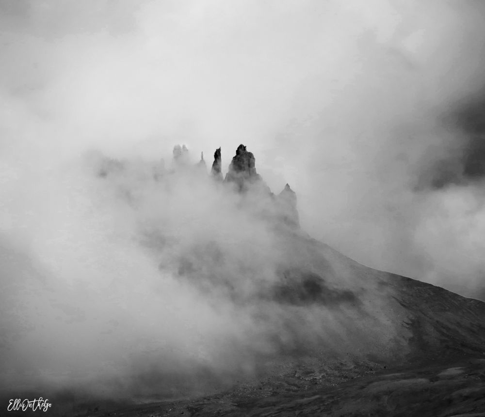 The image shows a dramatic, black-and-white scene of a mountain partially hidden in thick mist. Dark, jagged basalt peaks rise sharply from its summit, piercing through the swirling fog like the spires of a fortress. The surrounding slopes fade into the haze, giving the impression that the peaks are floating in a mysterious, cloud-filled void.