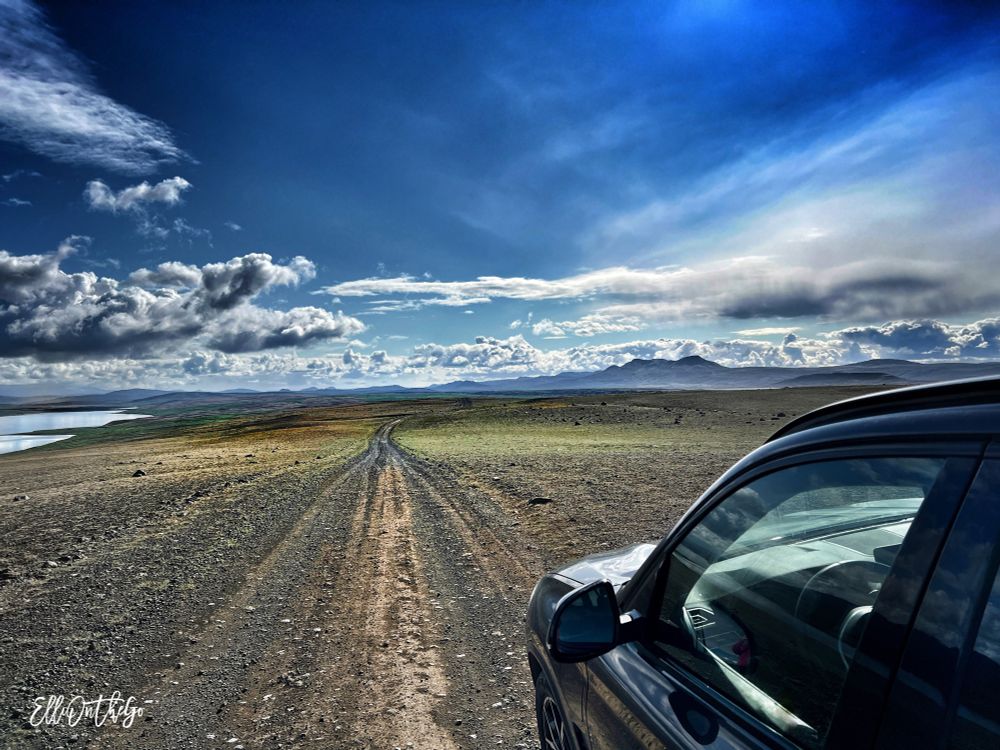 A wide, open landscape stretches into the distance under a dramatic sky. A rough dirt track runs straight ahead through a barren, rocky plain, curving slightly toward the horizon. On the right side of the image, the front half of a dark-colored SUV is visible. The land is mostly brown and grey, but there are subtle hints of green in the distance. To the far left, a narrow lake reflects the sky. Above, the sky is vast, filled with textured clouds and sunlight breaking through, creating a powerful sense of space, freedom, and solitude.