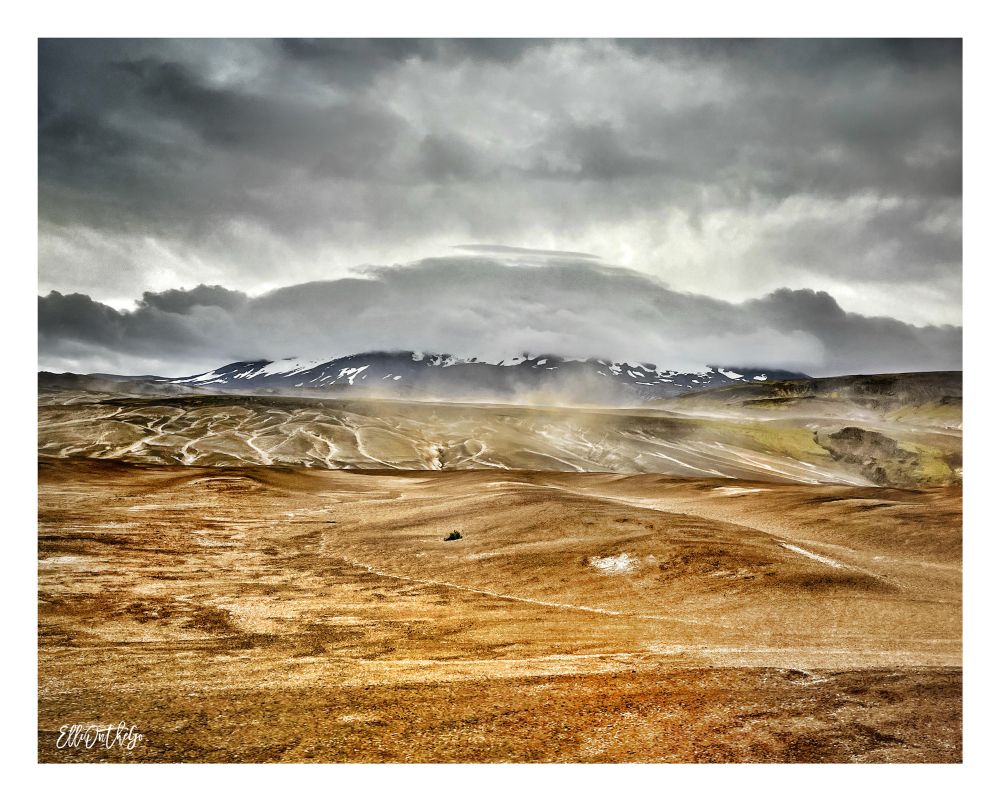 You’re standing in a vast, otherworldly landscape - raw, wind-swept, and breathtakingly stark. In the distance rises the volcano Hekla, one of Iceland’s most infamous peaks, its upper slopes brushed with streaks of snow like icing dragged by a hurried hand. But today, Hekla hides its crown in thick, swirling clouds, as if keeping ancient secrets tucked beneath a heavy gray sky.

Beneath the volcano, something extraordinary unfolds: a sandstorm dances across the barren plain. It doesn’t roar, but hisses and whispers as gritty gusts sweep over the ochre earth. Fine dust lifts in delicate, chaotic ribbons, painting the scene with motion and mystery. The ground is a mix of reddish-brown and pale , crisscrossed with natural veins and patterns etched by time and wind. In places, a hint of green moss clings to the surface - stubborn life in an otherwise harsh and barren world.

Despite the storm, there’s a strange calmness here - an eerie, lunar silence broken only by the unseen force of air carving through the silence. It’s not just nature - it’s raw drama, carved in volcanic stone and shaped by Arctic wind.

This is stunning in the truest sense: wild, untamed beauty that leaves you a little breathless.