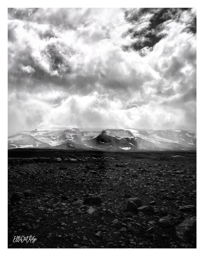 A dramatic black and white photo shows a stark volcanic landscape in Iceland. In the foreground, rough, dark lava rocks cover the ground. In the background, snow patches cling to the slopes of a wide, eroded crater - a long-extinct volcano near the Langjökull glacier. Thick clouds dominate the sky above, with light breaking through in places, casting a moody, almost otherworldly atmosphere across the scene. The image conveys solitude, timelessness, and raw natural power.