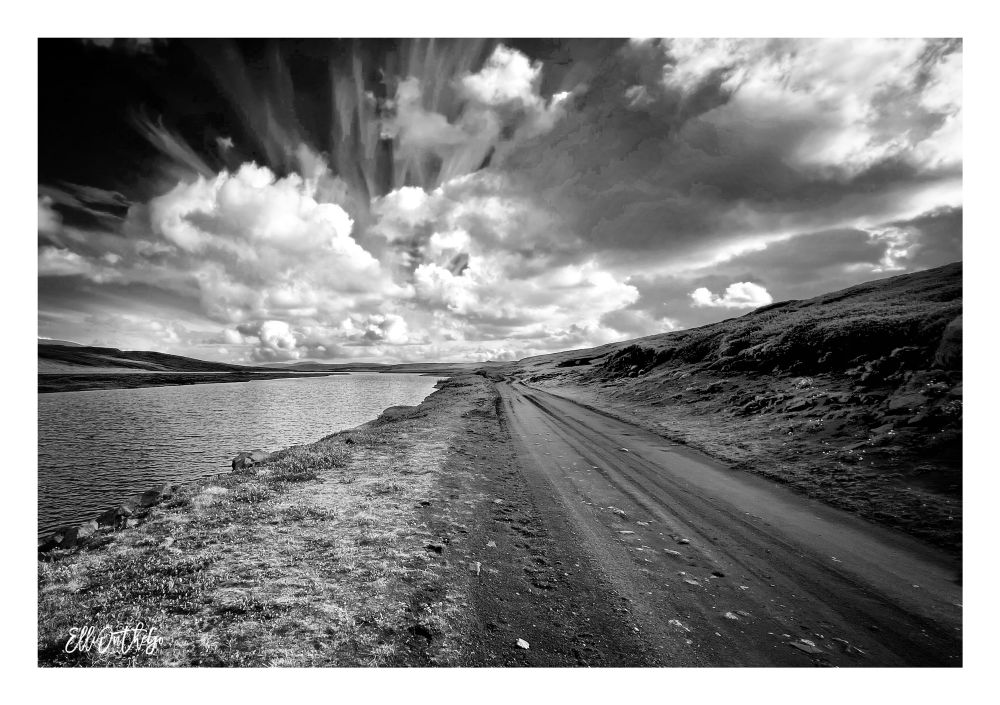 You’re standing beside a rough, unpaved track called Brúarvegur, deep in the eastern highlands of Iceland. The photo is in black and white, which brings out the stark contrasts of the landscape. To your left lies Þverávatn, a calm highland lake, its surface smooth and dark, quietly mirroring the sky.

To the right, the dirt road stretches ahead, marked by faint tire tracks, leading into the wide, open distance. Low hills flank the scene - not steep or mountainous, but gently rolling and mostly covered in moss, giving the terrain a soft, textured appearance, even in monochrome.

Above it all, the sky is vast and dramatic. Towering clouds spread across the horizon - some dense and heavy, others streaked and windswept - creating a sense of depth and movement. The black-and-white tones emphasize their shape and energy, turning the sky into something almost sculptural.

This is a scene of solitude and quiet strength - a raw, open place where the elements shape everything, and where the road invites you to disappear into Iceland’s untouched highland wilderness.