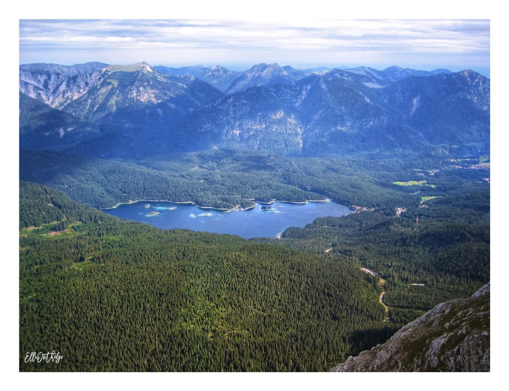 This photo shows a breathtaking view from high above, taken from the cable car on the way up to Germany’s highest mountain, the Zugspitze. Below, nestled among dense dark green forests, lies a large, shimmering lake - the Eibsee. The water appears deep blue with patches of turquoise near the shore and around small islands dotting the lake’s surface. Surrounding the lake, thick forests stretch into the distance, rising up into steep mountain slopes. In the background, layers of rugged alpine peaks fill the horizon under a sky filled with soft, streaky clouds.