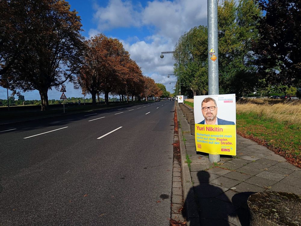 Straße mit vierspuriger Fahrbahn, rechts und links Gehweg, davon einer "Fußweg für Radfahrer frei". 
Rechts ein Wahlplakat der FDP mit dem Text "Sicherheit erreicht man nicht auf dem Papier, sondern auf der Straße"