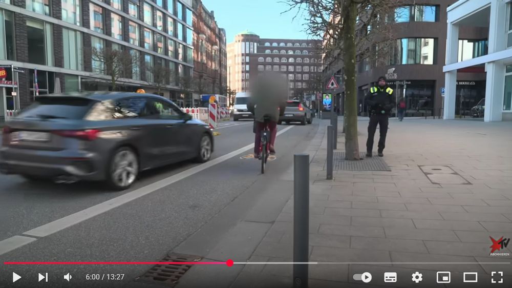 Links ein Auto auf der Fahrbahn, direkt daneben ein verpixelter Radfahrer auf dem Radweg. Rechts auf dem Gehweg ein Polizist.