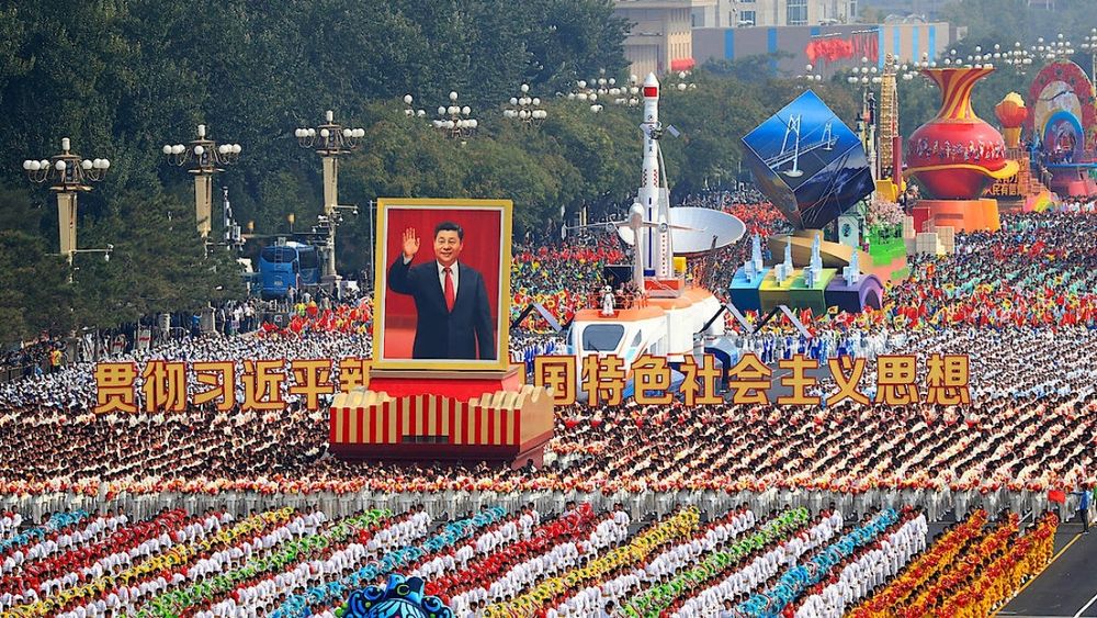 Parade mit mehreren tausenden Menschen in bunter Kleidung, in der Mitte ein riesiges Portrait von Mao Zedong 