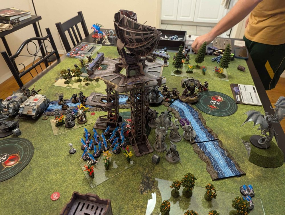 A warhammer game featuring Tyranids vs Space Marines with a large SATCOM tower in the middle made of cardboard. Painted river terrain flows down the center and around the tower, and there are a lot of woodland terrain pieces on the table. 