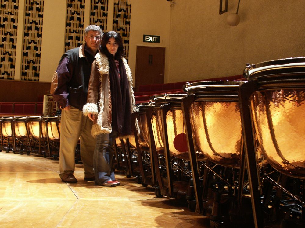A dramatic, colour photo of Jonathan and Evelyn, stood beside a bank of Timpani, with 11 in shot, on stage at the Liverpool Philharmonic Hall. The bowls are bright, shiny, hammered textured copper, reflecting the light beautifully. Both Jonathan and Evelyn are wearing several layers, with their coats on.