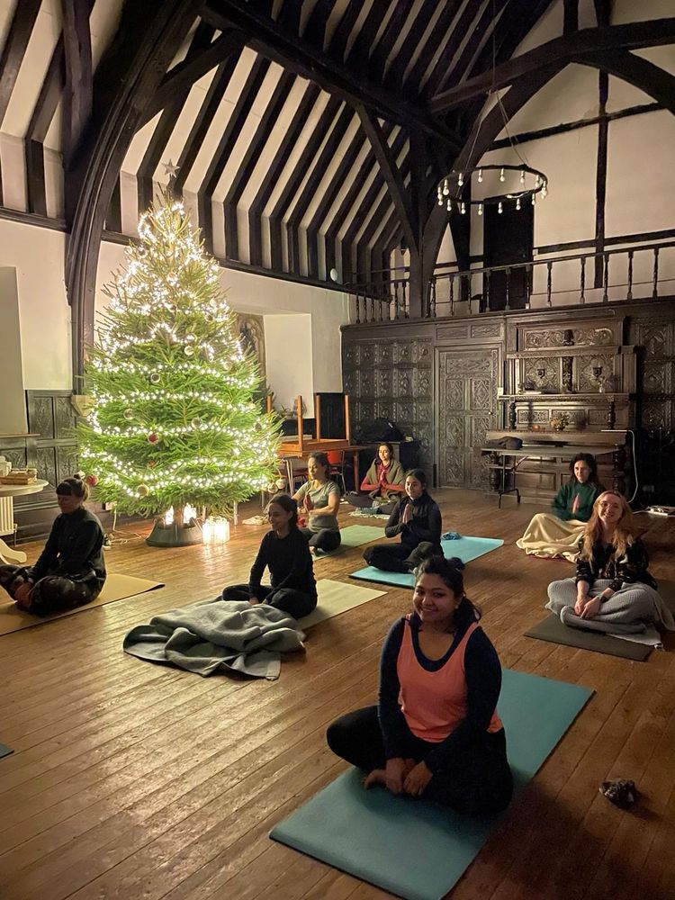 A very tall Christmas tree lights a large hall with a vaulted ceiling, bathing it it a white lights. Yoga mats are laid out on the floor, each with someone seated on it. 