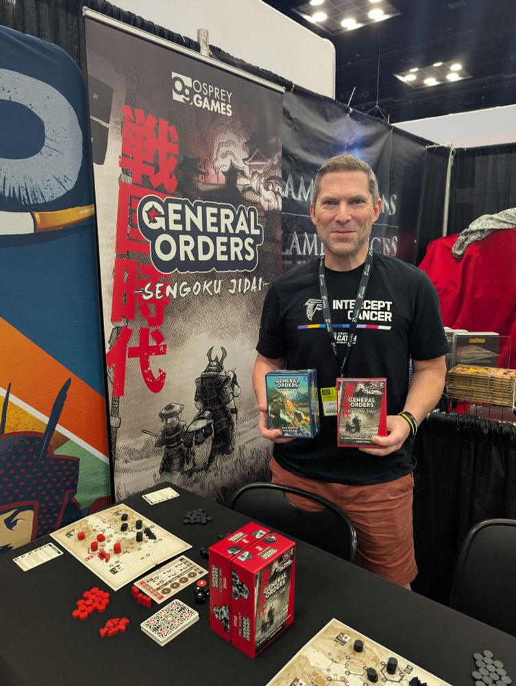A photo of co-designer David Thompson smiling and holding the boxes for General Orders: World War II and General Orders: Sengoku Jidai on the Osprey Games stand at Gen Con 2025.