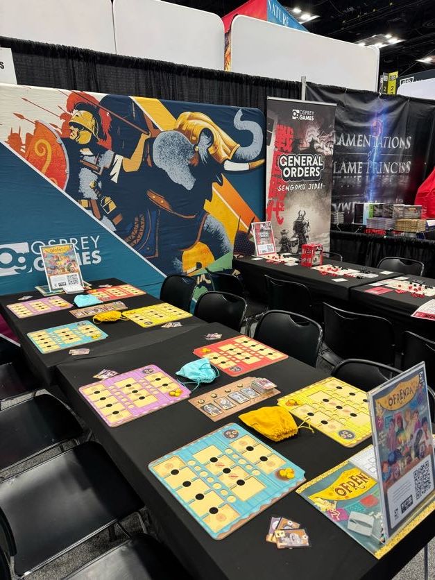 A photo of the Osprey Games stand at Gen Con 2025. Tables are set up with demo copies of the board games Ofrenda and General Orders: Sengoku Jidai, in front of banners with large, colourful art from various Osprey games.