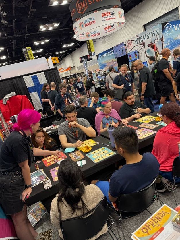 A photo of the stand full of people sat trying the demo games while a member of the Osprey Games team teaches them how to play.