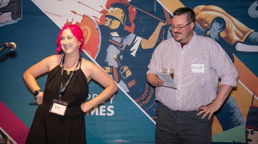 A photo of two members of the Osprey Games team stood smiling in front of a banner of illustrations from various board games.