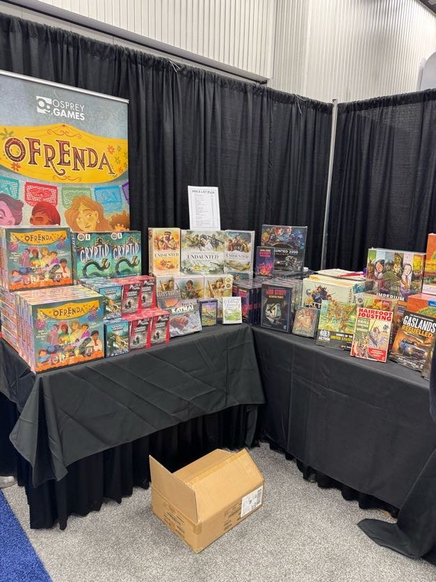 A close-up of the shop on stand, with two tables covered in black tablecloths and stacks of board games and wargame rulebooks, including Ofrenda, Cryptid, General Orders, Undaunted, Imperium, Bolt Action, Hairfoot Jousting and Gaslands. Behind is a banner for Ofrenda.