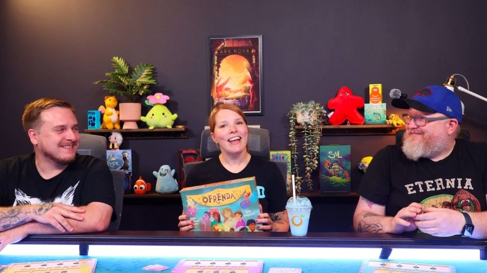 A screengrab from the official Ofrenda playthrough from Foster The Meeple on YouTube, showing the three hosts smiling and holding a copy of the game in front of shelves with various board games.