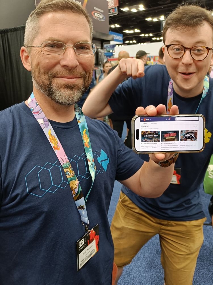 A photo of board game designer David Thompson at the Gen Con convention in 2024, wearing a t-shirt for Undaunted 2200: Callisto, holding a phone that shows Undaunted 2200: Callisto and War Story: Occupied France at the top of the show's hotness tracker on BoardGameGeek, while a member of the Osprey Games team points at it and smiles.