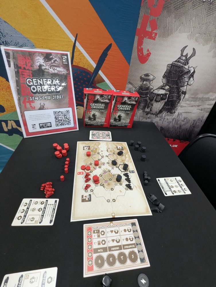 A photo of General Orders: Sengoku Jidai being demoed at Gen Con 2025. A game board is set up with various wooden pieces in red and black, surrounded by a support board and reference cards. In the background is a pair of game boxes and a banner showing off the cover art of medieval Japanese soldiers in a black-and-white style.