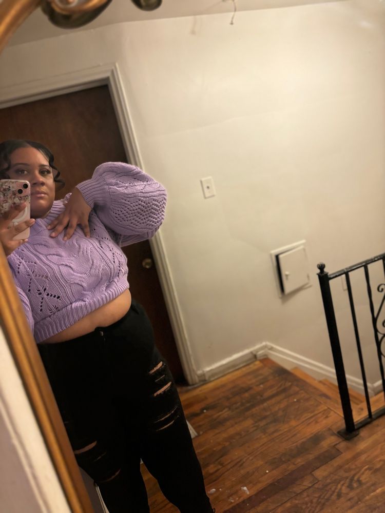 Beautiful black woman with black jeans , black boots, and purple crop top sweater standing in front of mirror. 