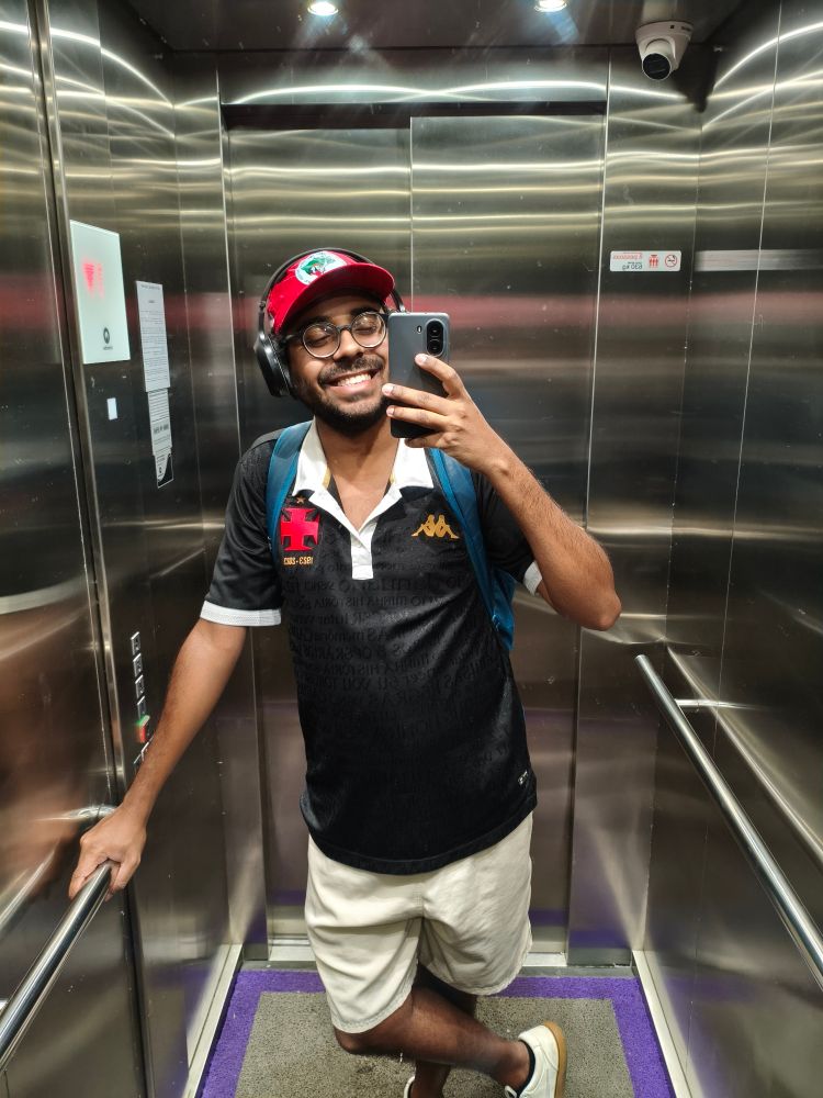 Eu, homem negro de sorriso maravilhoso, vestindo boné do mst, segurando celular na altura do rosto, camisa preta do vasco e short creme dentro de um elevador 