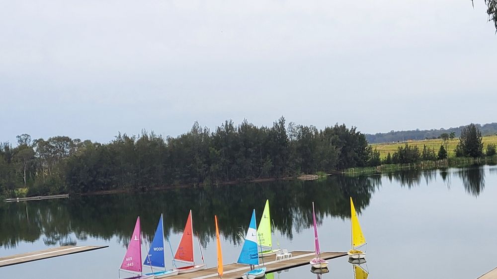 Pastel coloured boats and sails on blue wayerway with green trees on the horizon and blue sky. Pink, purple, green, blue, orange, yellow.