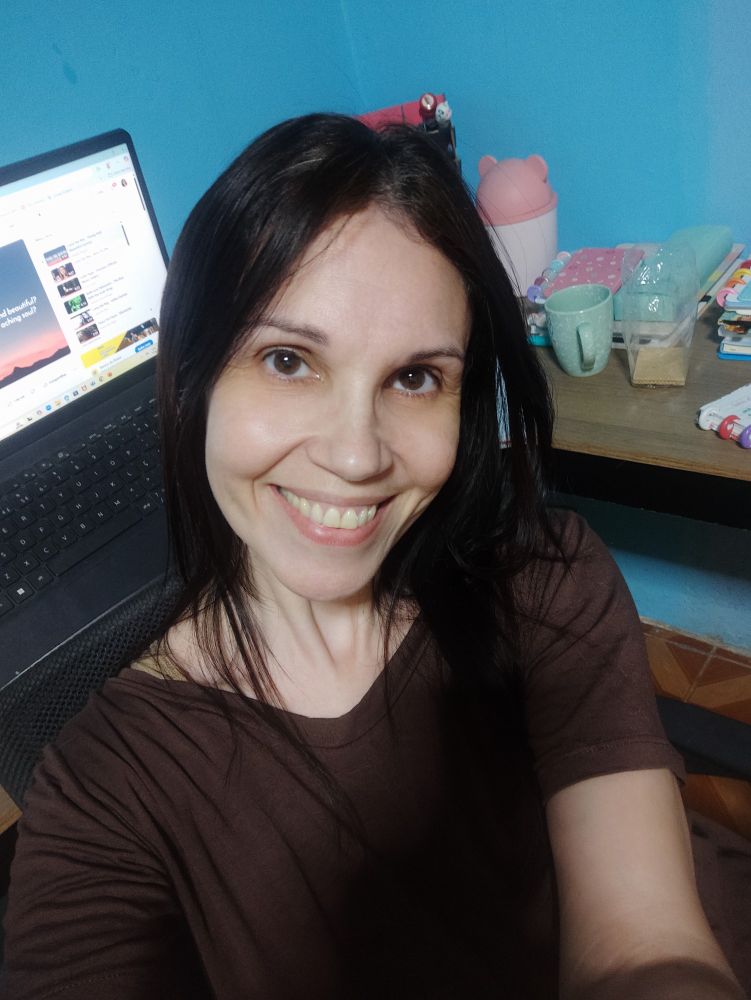 Mulher sorrindo (eu) com um laptop e uma mesa de trabalho ao fundo. 