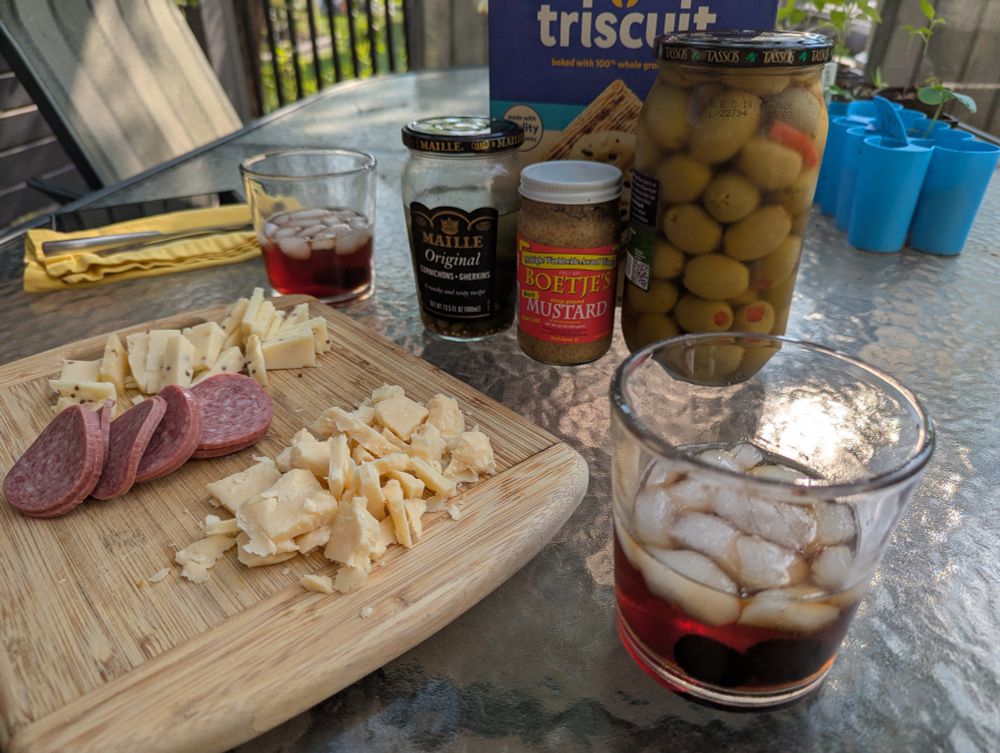 A lovely charcuterie board with two kinds of cheese and sliced salami sitting in front of jars of pickles, mustard, and green olives. Also pictured are two rye Manhattans in rocks glasses. All are sitting on the table on our deck in the back yard. Yum!