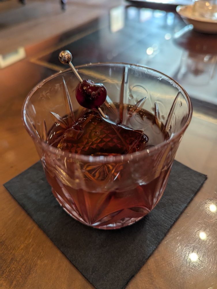 A very fancy glass holding a bourbon cocktail with a single square ice cube and a metal stirerer with a cherry on it.