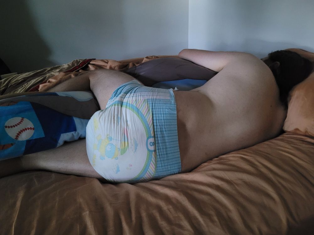Blankey is laying in bed with his back to the camera, arm and leg wrapped around his Brooks body pillow. His Teddy Ultra diaper is centered in the photo and is very wet