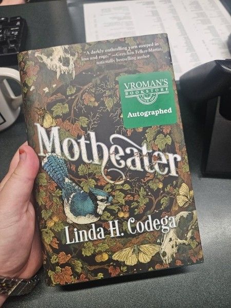 A white hand holding Motheater -- a book by Linda H. Codega -- with a sticker from Vroman's Bookstore that says "Autographed." The cover is dark and moody, a vortex of branches and dark green and brown leaves, with Cicadas, suckks, and moths surrounding it. There is a blue jay prominently drawn on the left hand side. The cover illustration is by Erica Williams.  