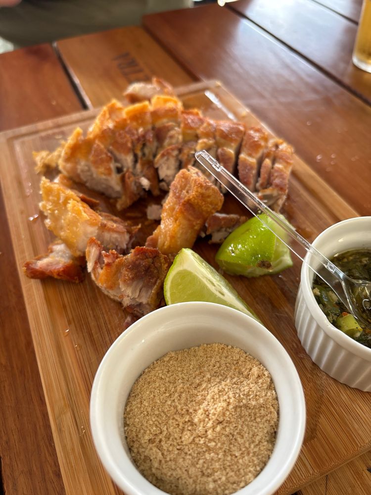 Tábua de madeira onde repousa torresmo de rolo cortado, fatias de limão e porções de farofa e chimichurri