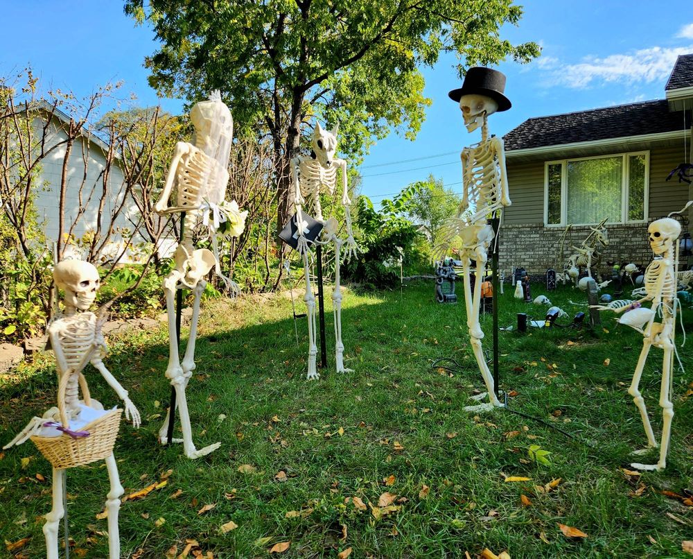 A werewolf skeleton officiates a wedding between a skeleton wearing a top hat and a skeleton wearing a veil. One smaller skeleton holds a basket of flower petals, and another smaller skeleton holds a pillow with rings tied to it.
