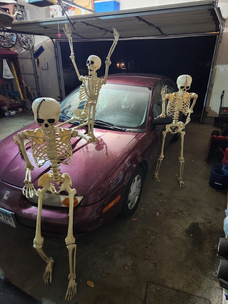 Three life-size skeletons are posed around my car. One is leaning on the front, a second is standing next to the driver's door, and the third is sitting on the hood with its arms in the air. Three more skeletons are inside the car. Two in the backseat, and one in the front passenger seat. They are all wearing their seatbelts.