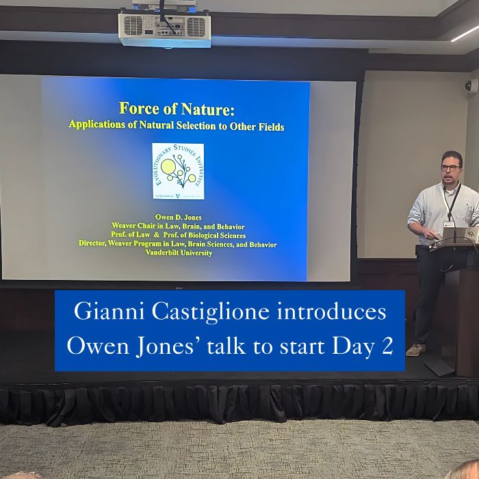 A man wearing a light blue button-down shirt and dark pants stands at a podium on the right side of the image, speaking in front of a projected slide that reads “Force of Nature: Applications of Natural Selection to Other Fields” by Owen D. Jones of Vanderbilt University. A blue caption at the bottom reads, “Gianni Castiglione introduces Owen Jones’ talk to start Day 2.”

