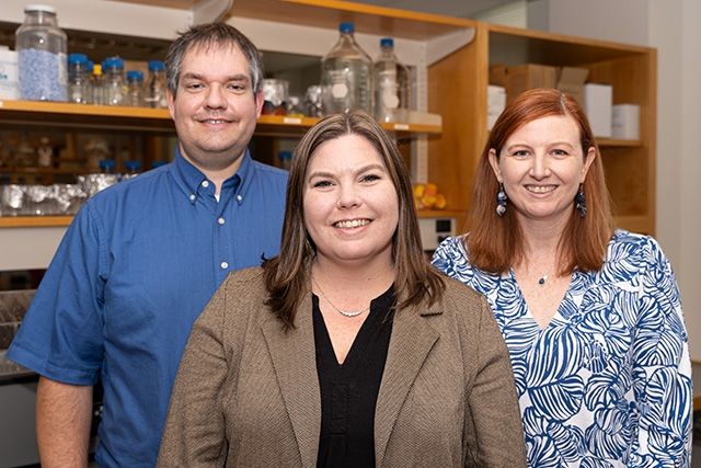 Three members of a research team are pictured in a laboratory. The man on the left wears a blue button- down shirt, while the two women beside nim, with brown and red hair, respectively, are dressed in professional attire, smiling at the camera. Laboratory shelves with various bottles and containers are visible in the background.