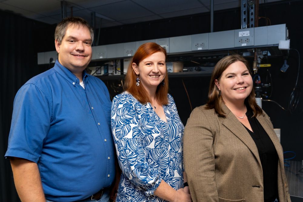 Three members of a research team are pictured in a laboratory. The man on the left wears a blue button- down shirt, the woman in the center has long red hair, wearing a blue and white patterned blouse. On the right stands another woman with brown hair, wearing a tan blazer over a black top. They stand in front of lab equipment and cables.