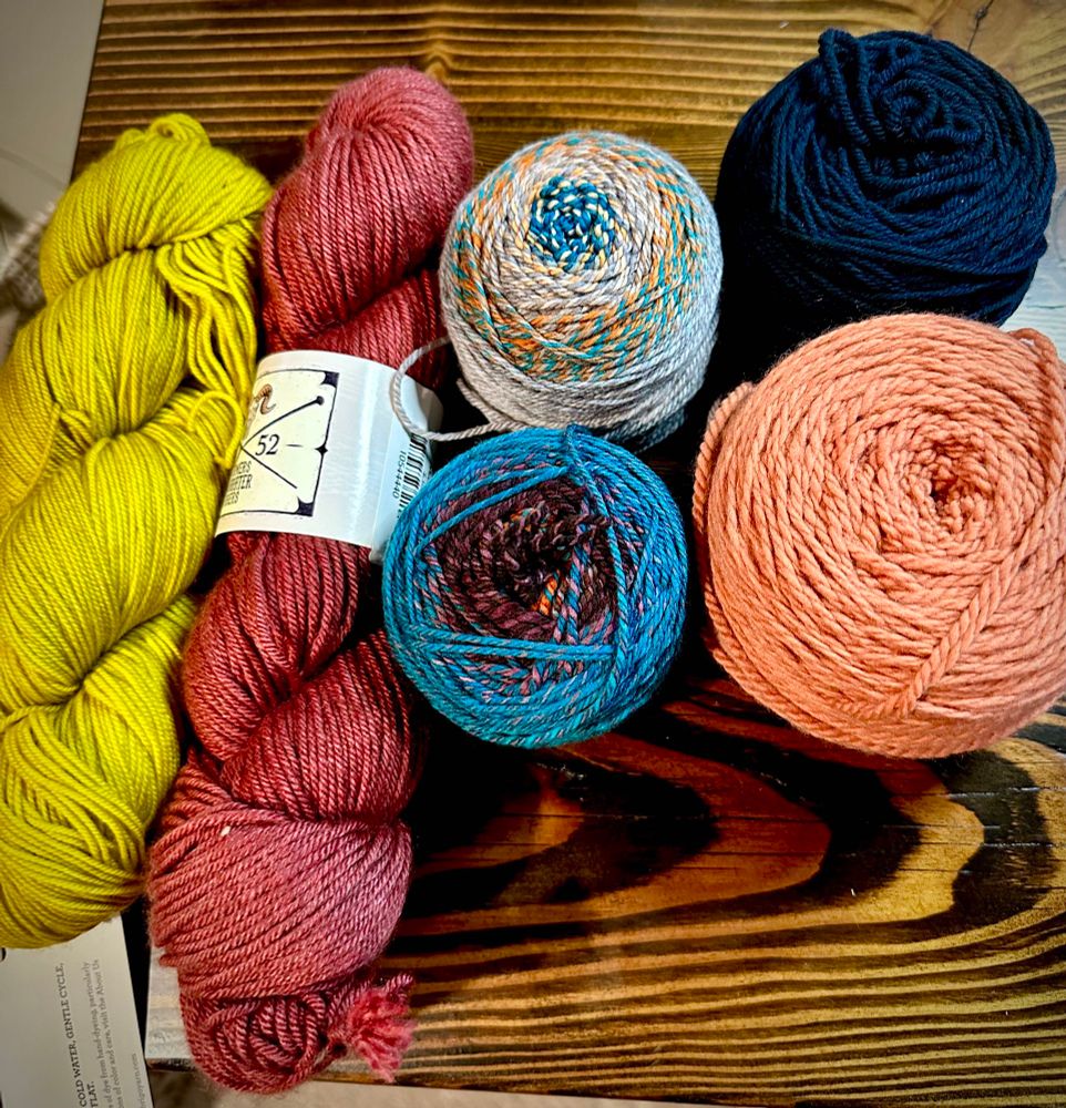 Six skeins of yarn, one yellow, one red, two Spincycle color shifting yarns, and orangey skein and a very dark blue skein all sitting in a dark wood table. 