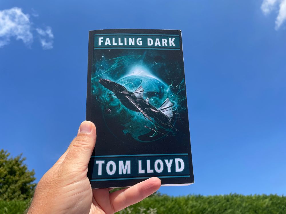 A hand holding up a paperback edition of Tom Lloyd’s “Falling Dark” against a clear blue sky.