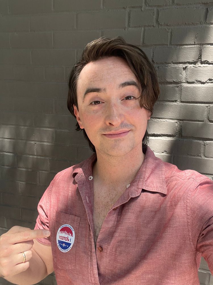 Me standing in front of a brick wall pointing to my voting sticker